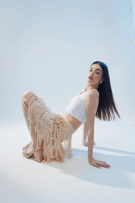 Young woman in a white top and beige fringed skirt poses on the floor in a bright studio. Concept of beauty, fashion, and modern style with soft natural lighting.