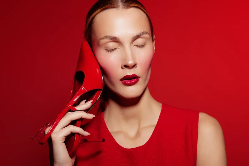 Woman with closed eyes holds red high heel shoe near face against red background. Fashion concept with matching red dress and lipstick, emphasizing style and elegance.