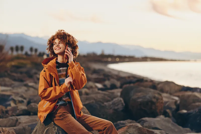 woman lifestyle outdoors at sunset, curly hair, orange jacket, smiling and relaxed while sitting on rocky beach, warm golden light, soft breeze, distant mountains, peaceful mood
