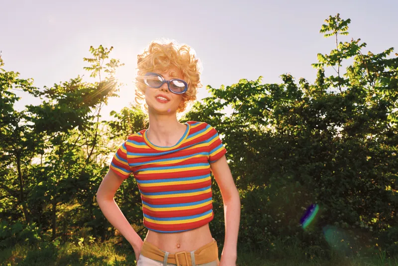 Cheerful woman outdoors, sunglasses and striped crop top, sunlit moment with a bright smile, casual pose under warm breeze, vibrant nature and retro film color mood
