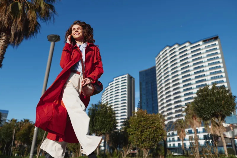 Young woman with curly hair wearing a red coat walks confidently in an urban park on a sunny day with modern buildings in the background and a joyful expression on her face.