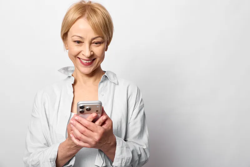 Elderly woman smiling in portrait indoors, holding smartphone, joyful expression, expression, technology for communication, casual lifestyle, white studio background