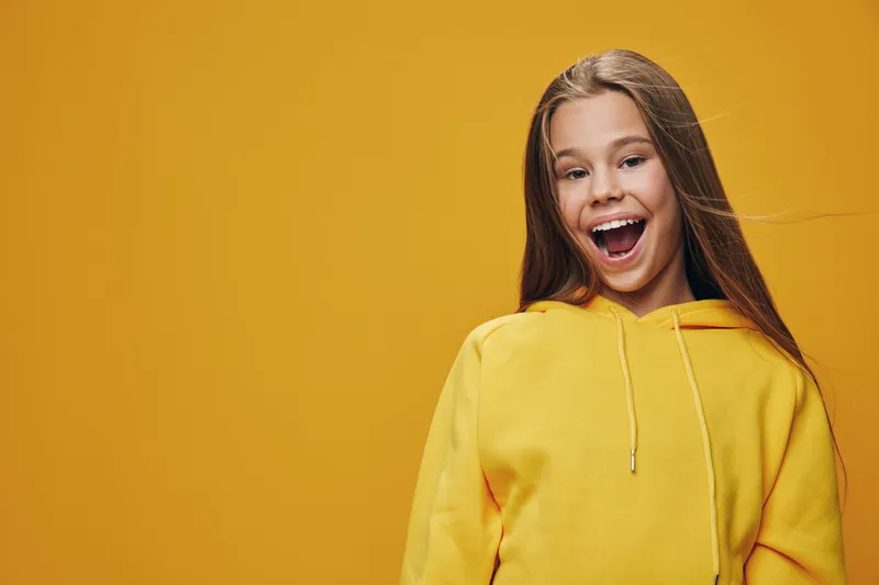Girl with long hair wearing yellow hoodie smiling and looking happy on orange background. Casual clothing, youth, positive emotion and lifestyle with cheerful expression.