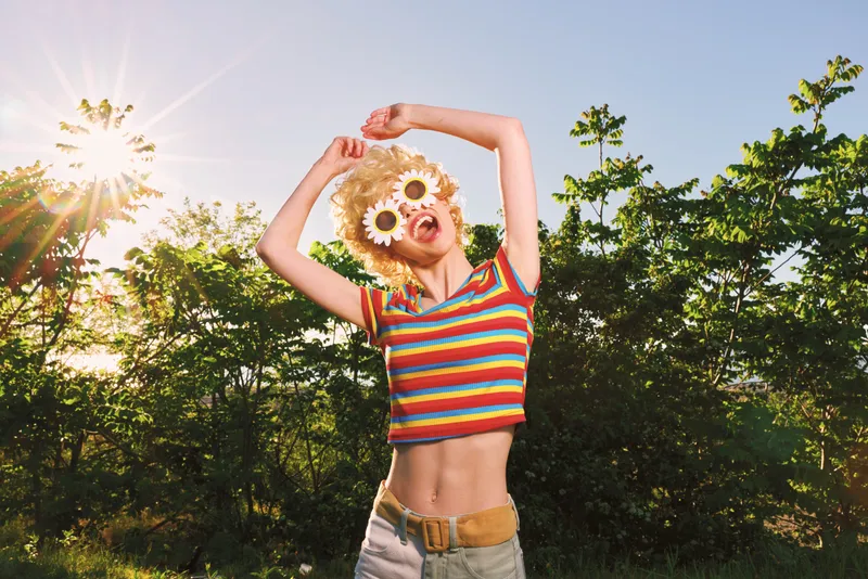 A cheerful woman in a striped shirt poses outdoors in bright sunlight, wearing sunflower sunglasses. Retro film color highlights a carefree summer lifestyle and playful fashion.