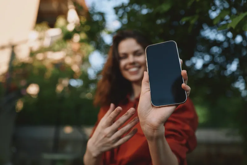 A cheerful woman in casual attire holds a smartphone for a selfie outdoors, smiling as she gestures toward the screen. A lifestyle moment captured in natural light