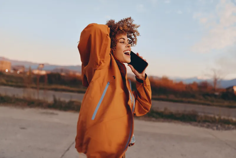 Woman lifestyle outdoors, orange raincoat with hood, laughing while calling on smartphone, standing on road at golden sunset, soft blurred landscape, warm evening mood, light breeze