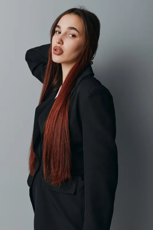 Young woman with long straight hair wearing black blazer posing with hand behind head against gray background. Fashion model with confident expression and stylish casual look in studio.