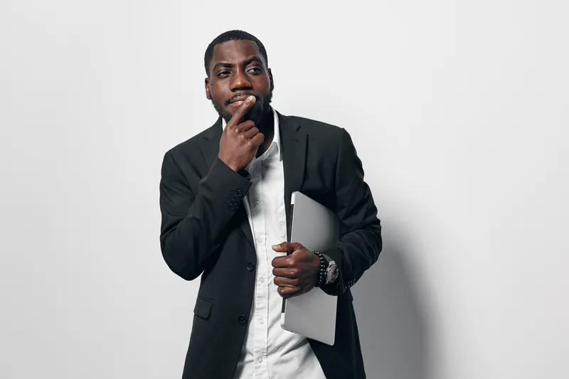 African American man with dark skin wearing black blazer and white shirt holding laptop and thinking with hand on chin in studio with white background.