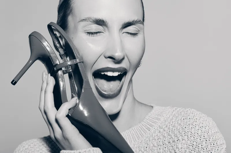 Happy Caucasian woman with pale skin holding a shiny high heel shoe near face laughing with closed eyes. Concept of fashion, joy, and footwear.