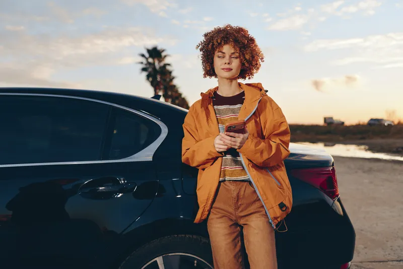Woman lifestyle portrait beside car, outdoors at sunset, curly haired woman in orange jacket holding phone, expression calm, golden sky, natural light, evening travel roadside atmosphere