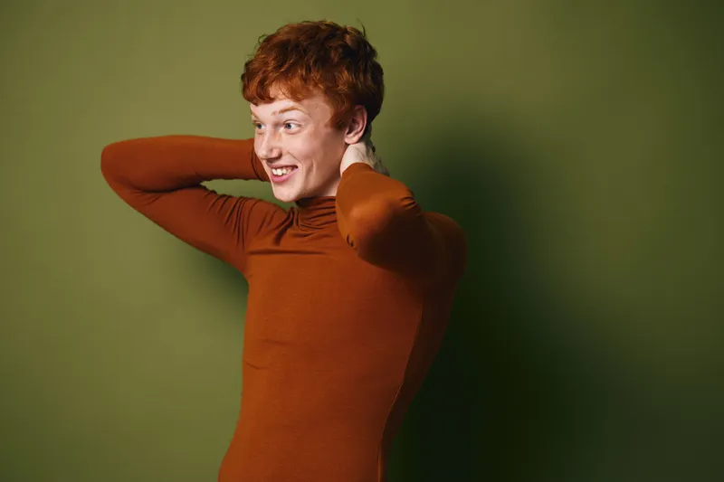 Portrait of smiling young woman with curly red hair wearing brown turtleneck posing with hands behind neck against green background in studio lighting.
