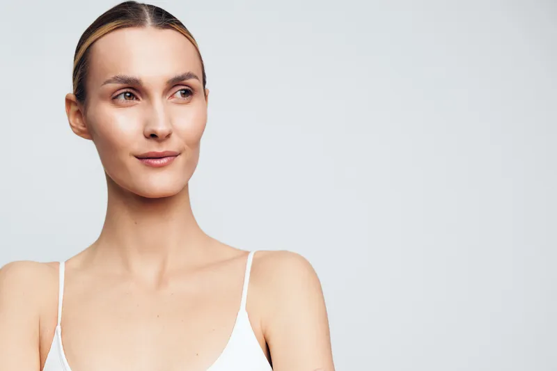 Woman portrait with clear skin and natural makeup in a studio, neutral background, soft lighting, calm expression, minimal styling, clean composition, conveying wellness and beauty