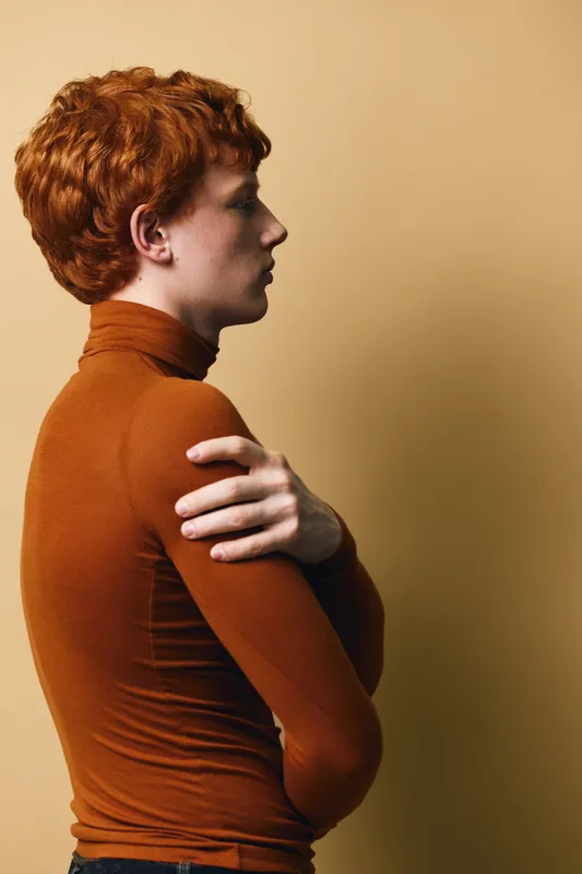 Redhead person in burnt orange turtleneck standing sideways against beige background with crossed arms, profile view, casual style and calm expression for portrait or fashion concept.