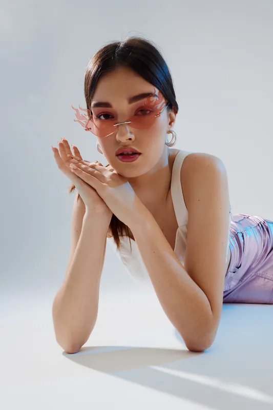 Young woman wearing pink sunglasses poses with hands near face in a white tank top and pastel skirt. Studio shot with soft lighting. Concept of beauty, fashion, and style.