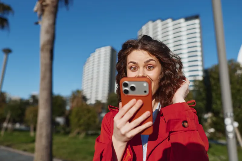 A young woman with curly hair in a red jacket looks surprised while holding a smartphone outdoors on a sunny day with urban buildings in the background.