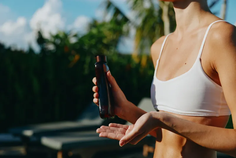 A fit person in a white crop top applies lotion from a dark bottle outdoors, sunlight beams on a lush garden background. Lifestyle composition highlighting skincare routine, outdoor wellness, and