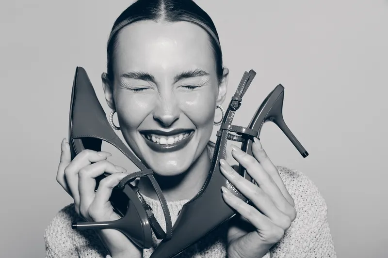 Happy white woman with fair skin holding two high heel shoes near her face smiling with closed eyes in studio. Concept of fashion and joy.