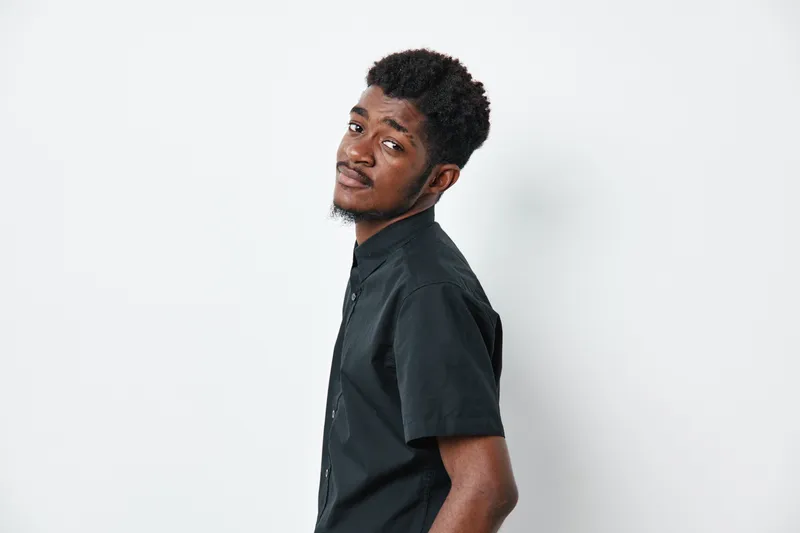 Young African American man with dark skin and curly hair wearing black shirt standing against white background looking at camera with confident expression.
