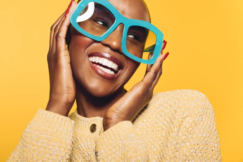 Happy smiling black woman with dark skin and short hair wearing large blue glasses and yellow sweater on bright yellow background. Concept of joy, fashion, and beauty.