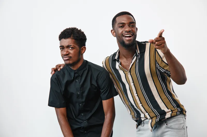 Two african men with dark skin posing together in casual clothes on white background. One man wears black shirt, other striped shirt, smiling and pointing with hand.
