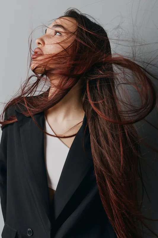 Woman with long hair flowing in the wind wearing black blazer and white top looking sideways on gray background. Fashion portrait, hairstyle movement and casual outfit with natural expression.