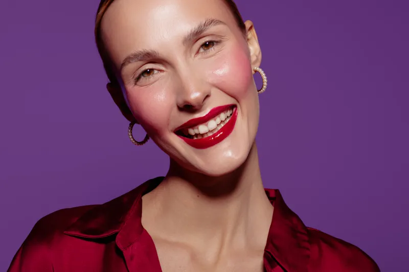 Smiling woman with red lipstick and hoop earrings poses against purple background. Close-up portrait with shiny skin and red satin blouse. Concept of beauty, fashion, and confidence.