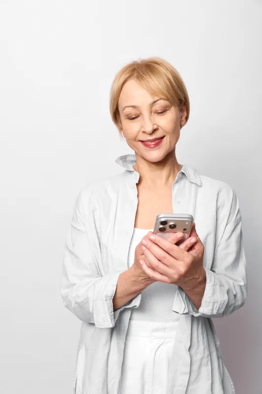 Senior woman holding smartphone and smiling, studio portrait on white background, natural communication concept, technology lifestyle, calm confident mood, soft light.