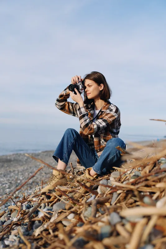 Woman photographer sitting on driftwood by the sea, wearing plaid shirt and jeans, aiming a camera toward the horizon for a coastal outdoor photography session.