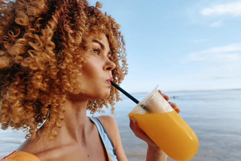 Curly woman drinks fresh orange juice from plastic cup with straw on beach, looks thoughtfully at sea, enjoys healthy tropical beverage and sunny summer day outdoors.