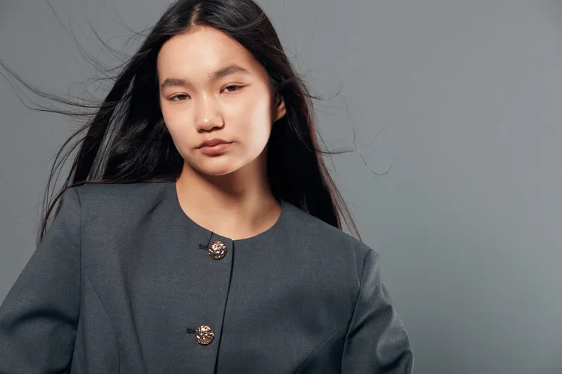 Young woman with long black hair wears grey coat with decorative buttons standing against neutral grey background in studio, looking confidently at camera with natural expression.