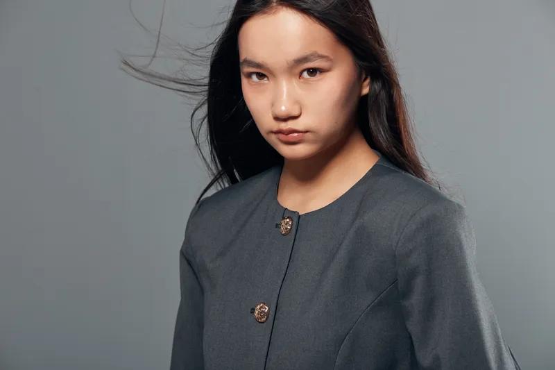 Young woman with long dark hair wearing a gray blouse with decorative buttons, looking serious and confident in a studio setting with plain gray background.