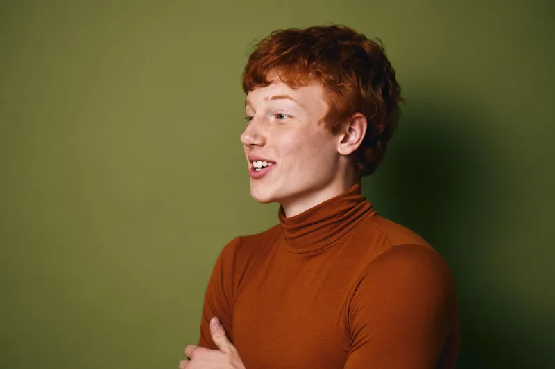 Young man with red hair and freckles wearing a brown turtleneck sweater smiling and looking sideways against green background in studio portrait shot.
