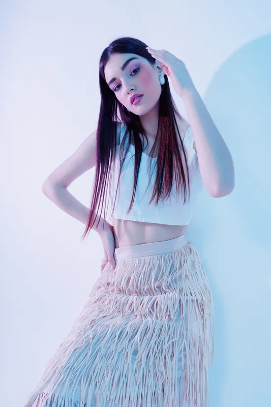 Young woman with long dark hair poses in a white crop top and fringed skirt. Soft lighting highlights her beauty and fashion style on a plain light background.