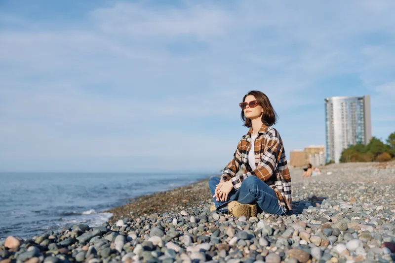 Casual person sits on a pebbled beach by the sea, wearing a plaid shirt and sunglasses, enjoying the breeze, distant buildings on the shore, and a calm sunny coastal scene with open horizon.