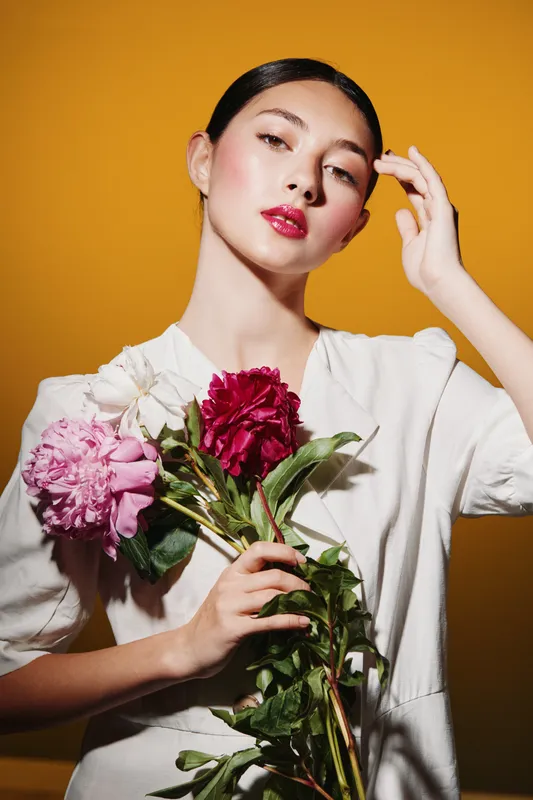 Young woman with sleek hairstyle poses confidently holding vibrant flowers against yellow background. Her red glossy lips and smooth skin highlight creative beauty concept and fashion style.