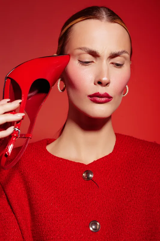 Woman in red sweater holds red high heel shoe near face with closed eyes and serious expression. Studio portrait on red background. Concept of fashion, style, and bold color contrast.