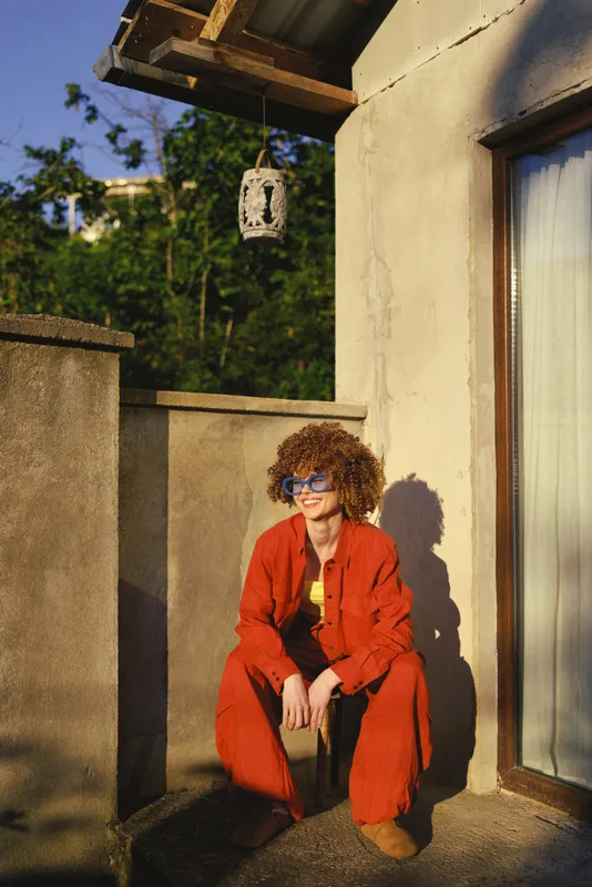 A woman with curly hair in a red jumpsuit sits on a sunlit balcony, relaxed and smiling, captured in a casual urban lifestyle moment outdoors, bright and warm