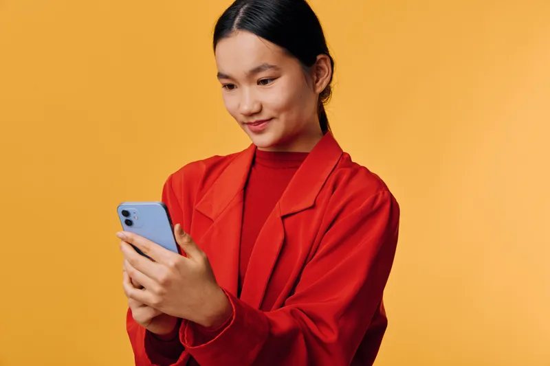 Young woman in red jacket using smartphone on orange background for communication, social media, technology and online connection with smile and casual outfit.