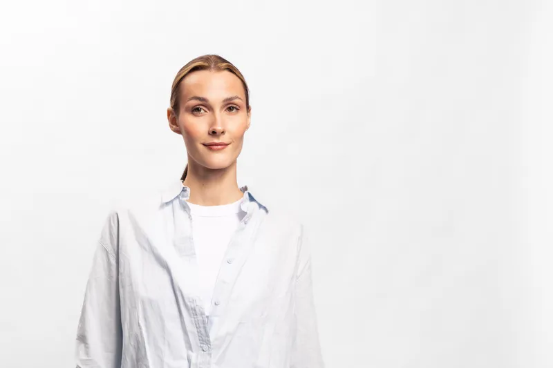Confident young woman with blonde hair wearing a white shirt stands against white background. Studio portrait with natural makeup and casual style. Concept of simplicity and self-assurance.
