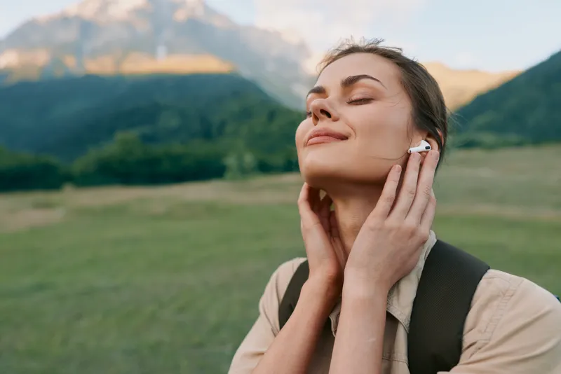 A woman enjoys nature outdoors with a calm, uplifted expression, listening to music. She wears a backpack in a grassy field with mountains in the background.