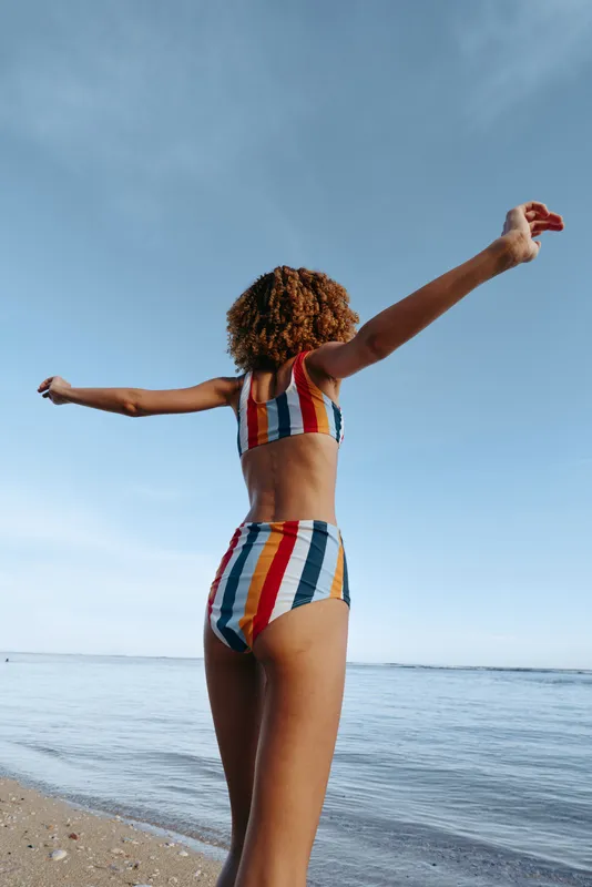 Woman, beach, ocean, swimsuit, curly hair, summer sunlight, freedom, standing with arms outstretched, seaside waves, relaxed vacation, clear sky