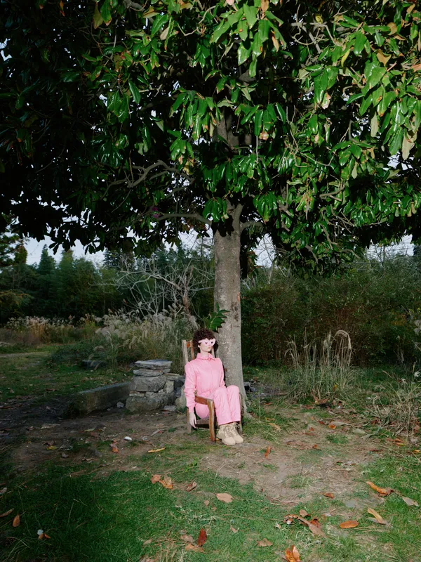 Woman, pink dress, sitting outdoors under a large tree in a garden, with fallen leaves and rustic rocks nearby, capturing a tranquil moment of solitude and nature.
