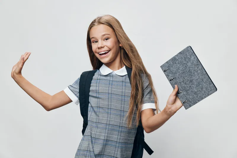 Girl student with backpack holding notebook smiling and waving hand in casual clothes on gray background for education and school concept.