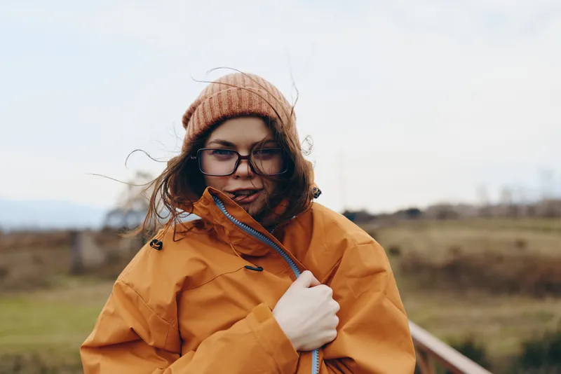 Woman in winter outdoor jacket and glasses with beanie smiles holding zipper, warm cold weather clothing in nature, wind and blurred landscape behind, lifestyle concept for travel