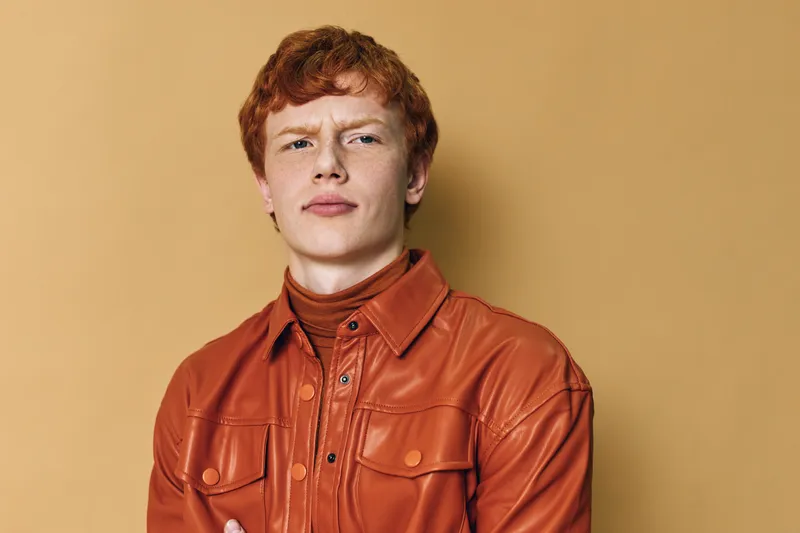 Young man with red hair wearing a brown leather jacket and turtleneck posing with crossed arms on plain background. Confident male portrait with casual fashion style and serious expression.