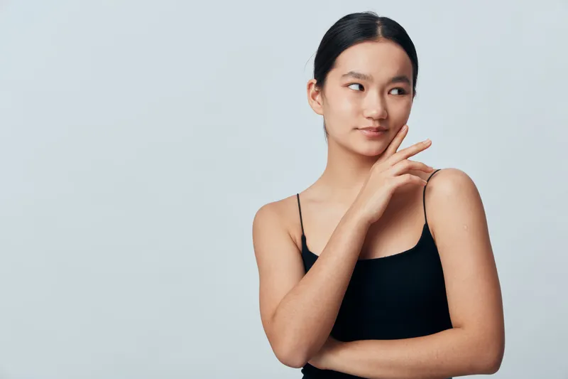 Young woman with dark hair and natural makeup wears black tank top standing against plain light background looking sideways with a subtle smile and relaxed arms pose.