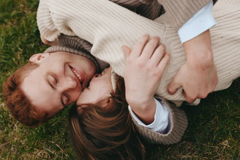 Two people lie on grass, embracing in a warm outdoor moment of couple romance and cuddling. Cozy sweaters glow in soft sunlight, conveying intimacy, happiness, and relaxed lifestyle in a peaceful