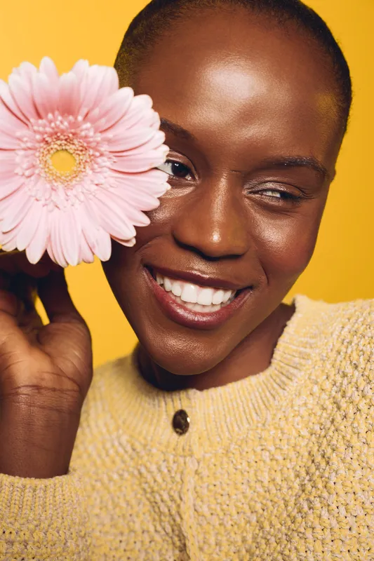 Smiling dark skinned African woman holds pink flower near face, wears yellow knitted sweater. Bright yellow background enhances joyful mood, concept of beauty, nature, and happiness.