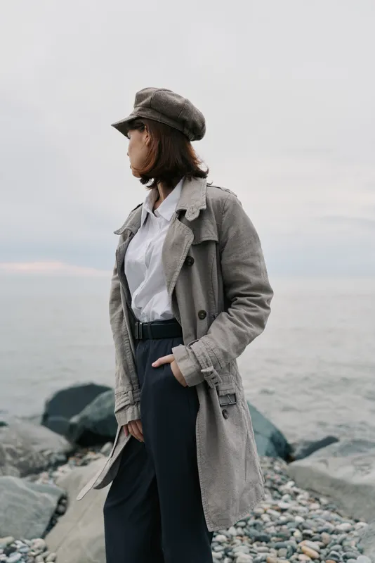 Young woman in trench coat and cap stands on rocky shore looking sideways with sea and cloudy sky in background. Concept for fashion, autumn, and outdoor lifestyle.