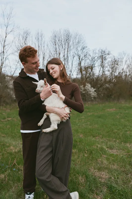 Cheerful couple outdoors, holding a goat kid, smiling beneath the sun, casual attire, open field, soft breeze, natural colors, lush grass, warm connection, relaxed pose, candid moment, authentic